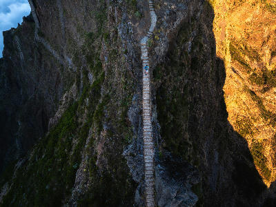 Aerial view of a hiker crossing a narrow stone path along the Pico do Arieiro–Pico Ruivo ridge, suspended above steep volcanic cliffs in Madeira.