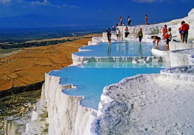 Visitors wading in turquoise thermal pools across the white travertine terraces of Pamukkale, Turkey, with the valley and mountains in the distance.