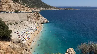 Elevated view of busy Kaputaş Beach, with rows of umbrellas and swimmers along a narrow sandy cove beside clear turquoise Mediterranean water and steep rocky cliffs.