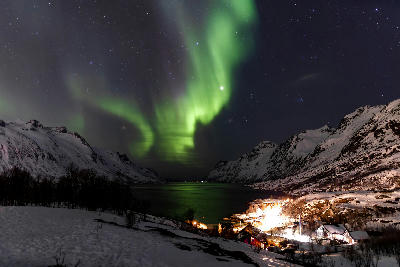 Green northern lights over Ersfjord fjord with snow-covered mountains and small illuminated village below