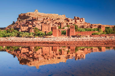 Aït Benhaddou fortified clay kasbah in Morocco reflected in water at sunrise with Atlas Mountain desert terrain and oasis vegetation