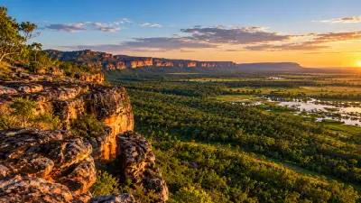 AI-generated panoramic landscape inspired by Kakadu National Park, showing golden sandstone cliffs, dense green woodland and reflective wetlands beneath a warm sunset sky in Australia's Northern Territory.