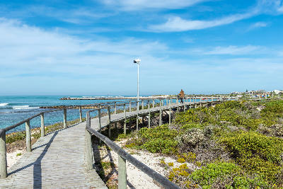 Boardwalk above coastal dunes and fynbos at Struisbaai with ocean and harbour views beneath blue skies in the Western Cape.