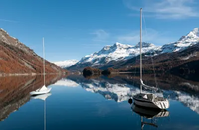 Two sailboats on a calm alpine lake reflecting snow-covered mountains and forested slopes at Lake Silvaplana near St. Moritz, Switzerland.