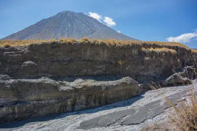 The symmetrical cone of Ol Doinyo Lengai towering above a dry volcanic gully with white carbonate mineral deposits and dark lava flows in the Great Rift Valley, Tanzania.