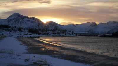 Snow covered Bøvær Beach Senja Norway with curved shoreline, small coastal houses and mountain range reflecting in calm fjord water at sunset.