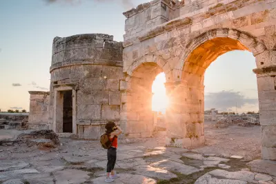 Visitor photographing the monumental stone arches of the Frontinus Gate in ancient Hierapolis as warm sunset light shines through.