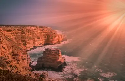 Island Rock stands offshore beneath layered sandstone cliffs as waves break around its base in warm evening light.