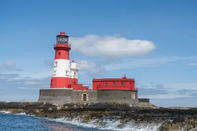 Red-and-white Longstone Lighthouse and adjoining buildings on a rocky island, with waves breaking along the North Sea coastline of the Farne Islands.
