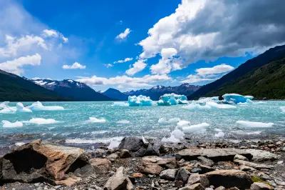 Icebergs floating on turquoise Lago Argentino with rocky shoreline and Andes mountains in Patagonia, Argentina.