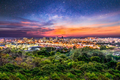 Panoramic view of Pretoria at dusk with illuminated city lights, surrounding greenbelt vegetation and a colourful star-filled sky in Gauteng, South Africa.