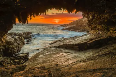 Orange sunset over the Southern Ocean framed by Admirals Arch, showing hanging limestone formations and waves breaking against the rocky Kangaroo Island coastline.