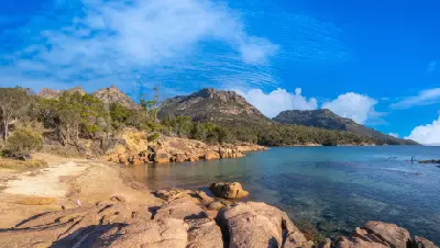 Pink granite rocks and clear sheltered water at Honeymoon Bay with the five peaks of The Hazards rising behind, Freycinet National Park, Tasmania.