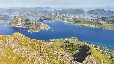 Aerial view from Stornappstinden toward Offersøykammen on Vestvågøya in the Lofoten Islands, showing fjords, green farmland and mountain ridges along the Norwegian coast.