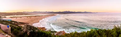 Panoramic coastal view of Lookout Beach in Plettenberg Bay with gentle surf, sandy shoreline and distant mountain ranges at sunrise.