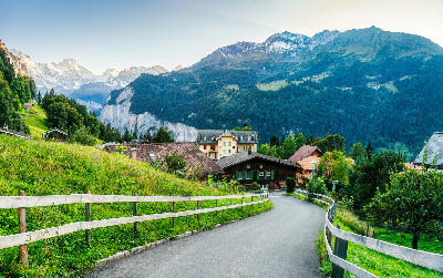Path leading through Wengen village with chalets and green meadows, overlooking Lauterbrunnen Valley and alpine mountains in the background, Switzerland.
