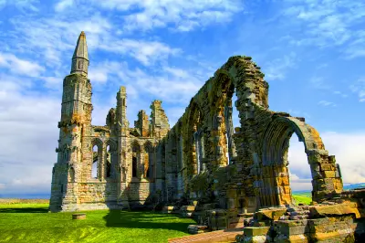 Ground-level view of Whitby Abbey's ruined Gothic arches, tall stone tower and weathered medieval walls beneath a blue sky in North Yorkshire, England.