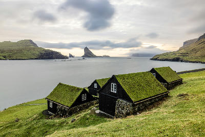 Black wooden houses with grass roofs in Bøur facing Tindhólmur islet and the Drangarnir sea stacks on Vágar Island, Faroe Islands.