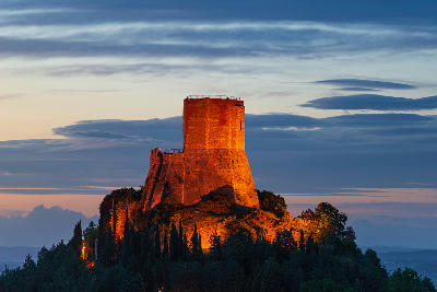Dusk view of Rocca di Tentennano above the Val d’Orcia, with the stone tower illuminated against a dramatic evening sky in Tuscany, Italy.