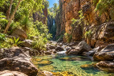 Clear creek pools wind between large boulders and towering red cliffs toward the waterfall at Emma Gorge.