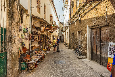 Narrow historic street in Stone Town, Zanzibar, with local craft shops, traditional baskets, and pedestrians exploring the old town.