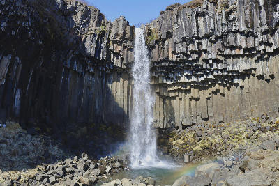 Öxarárfoss waterfall flowing over dark basalt cliffs into a rocky pool in Thingvellir National Park, Iceland, with a small rainbow visible in the mist.