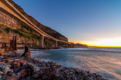 Low-angle coastal view beneath Sea Cliff Bridge at sunset, with the elevated roadway sweeping along sandstone cliffs above the Tasman Sea.