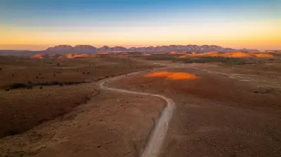Aerial sunset view of a winding dirt road through dry, rolling hills near Razorback Lookout, with golden ridgelines and distant mountains in South Australia's Flinders Ranges.