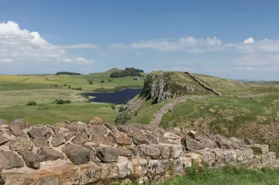 Hadrian's Wall crossing Highshield Crags above Crag Lough, with rocky cliffs, rolling green hills and blue summer sky in Northumberland, England.