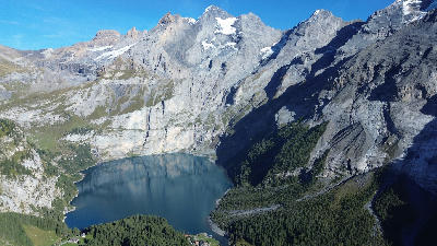 Aerial view of Oeschinensee, a mountain lake surrounded by steep cliffs, rocky slopes, and forest in the Bernese Alps above Kandersteg, Switzerland.