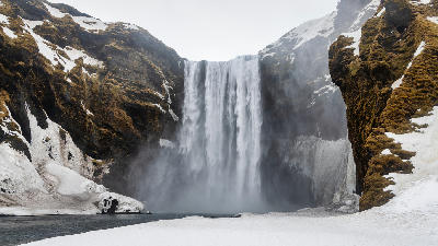 Skógafoss waterfall plunging from a cliff in winter, with snow, moss-covered rock, and overcast skies in southern Iceland.