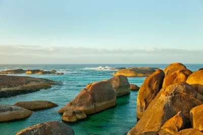 Large, smooth granite boulders are illuminated by warm morning side light in a calm turquoise bay, with ocean waves breaking in the background under a pale sky.