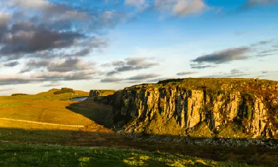 Sunset illuminating the steep rocky cliffs of Peel Crags in Northumberland, England, with grassland, distant hills and scattered clouds above the Hadrian's Wall landscape.