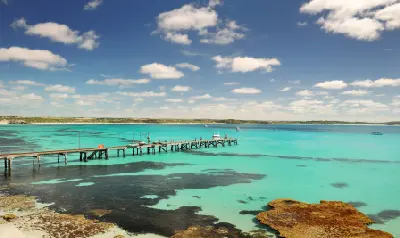 Old wooden jetty extending into turquoise coastal water on Kangaroo Island, South Australia, with submerged reefs, small boats and a partly cloudy sky.
