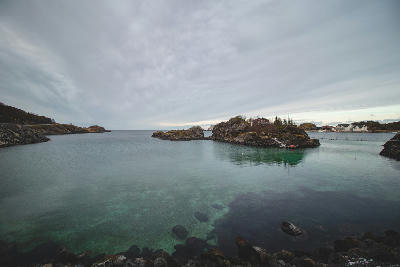 Calm turquoise harbour water with small rocky islets and village buildings at Hamn i Senja under overcast sky