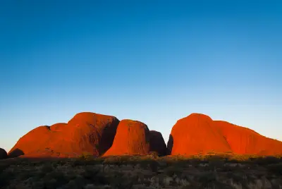Ground-level view of the sandstone domes of Kata Tjuta glowing red at sunset above dark desert vegetation.