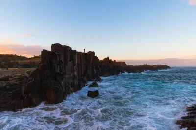 Silhouetted visitor stands atop the dramatic basalt columns of Bombo Headland as ocean waves break against the rocks at dusk.