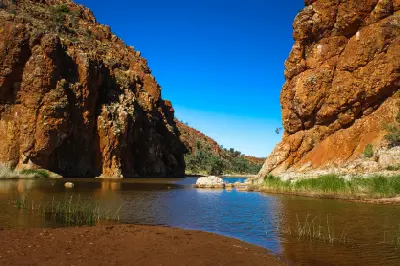 Sun-baked orange cliffs look down on a calm, deep waterhole at Glen Helen Gorge, with a dark, dry dirt bank in the foreground beneath a clear blue sky.