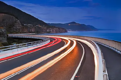 Long-exposure light trails sweep around the curves of Sea Cliff Bridge beside the Tasman Sea beneath a deep blue evening sky.