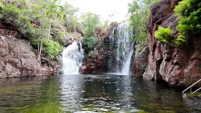 Twin waterfalls flowing into the natural swimming pool at Florence Falls, with sandstone cliffs, tropical vegetation and a metal access handrail in Litchfield National Park, Northern Territory, Australia.