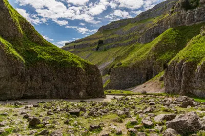 Ground-level view of Gordale Scar showing steep limestone cliffs, green grassy slopes and scattered rocks beneath a partly cloudy sky in North Yorkshire.