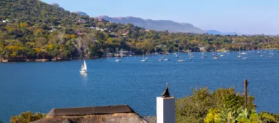 View across Hartbeespoort Dam with a sailboat on open water, numerous moored yachts near the shoreline, and the Magaliesberg mountains rising in the background.