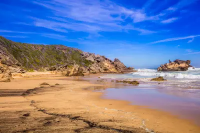 Beach at Brenton-on-Sea with rocky headlands, surf rolling onto golden sand and coastal vegetation beneath a blue sky near Knysna, South Africa.