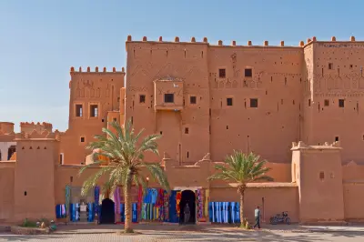 Front entrance of Kasbah Taourirt in Ouarzazate, Morocco, with adobe fortress walls, palm trees, and colorful market textiles.