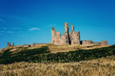 Southern view of Dunstanburgh Castle showing its ruined twin-towered gatehouse and long medieval stone walls above a grassy hillside beneath a clear blue sky.