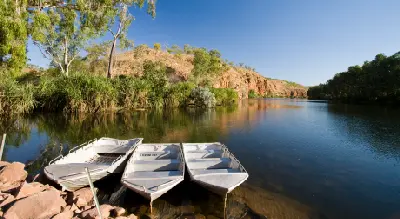 Small boats moored on the calm Chamberlain River beneath rugged sandstone cliffs and lush riverside vegetation in Chamberlain Gorge.