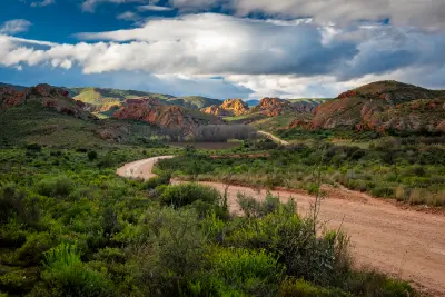Curving gravel road passing through red sandstone hills and Karoo vegetation at Red Stone Hills near Calitzdorp, with dramatic clouds and layered mountain landscapes in the Klein Karoo, South Africa.