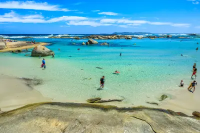Families and visitors dot the white sand and shallow crystal-clear green waters of a calm coastal swimming area.