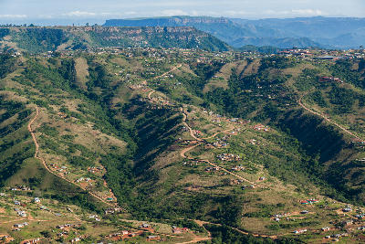Elevated view across the Valley of a Thousand Hills showing steep green valleys, winding rural roads, scattered homes and escarpment cliffs in the distance.