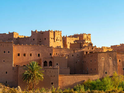 Kasbah Taourirt in Ouarzazate at sunrise with layered earthen walls, palm trees, and warm desert light.